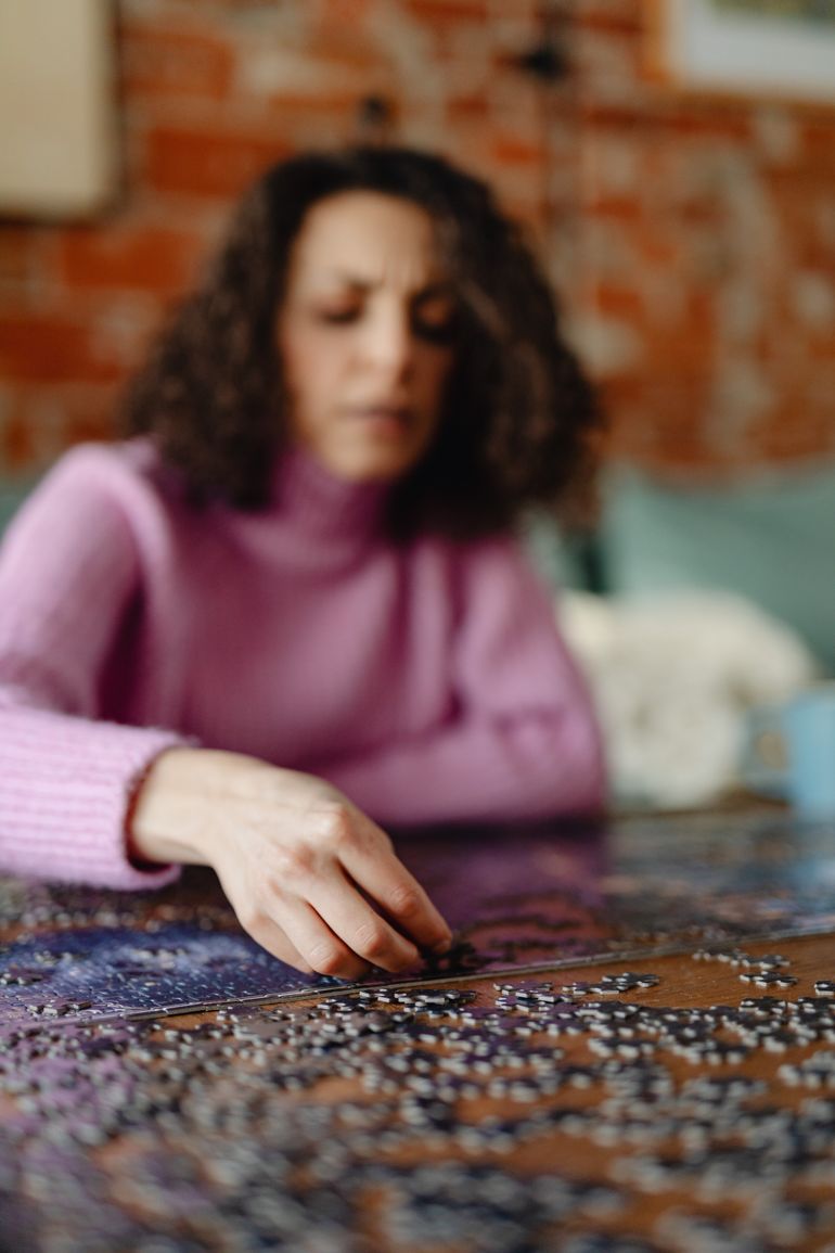 A woman wearing a pink sweater focuses on completing a jigsaw puzzle on a table.