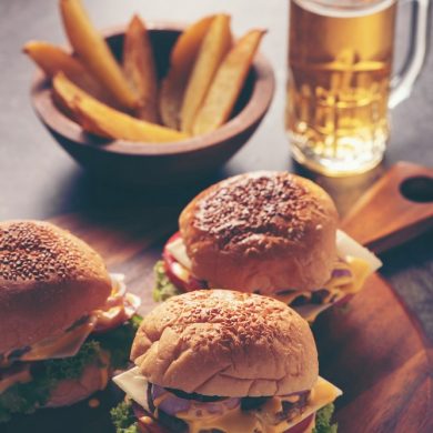 Three burgers sit on top of a wooden serving platter alongside French fry wedges and a beer