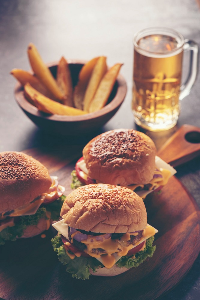 Three burgers sit on top of a wooden serving platter alongside French fry wedges and a beer